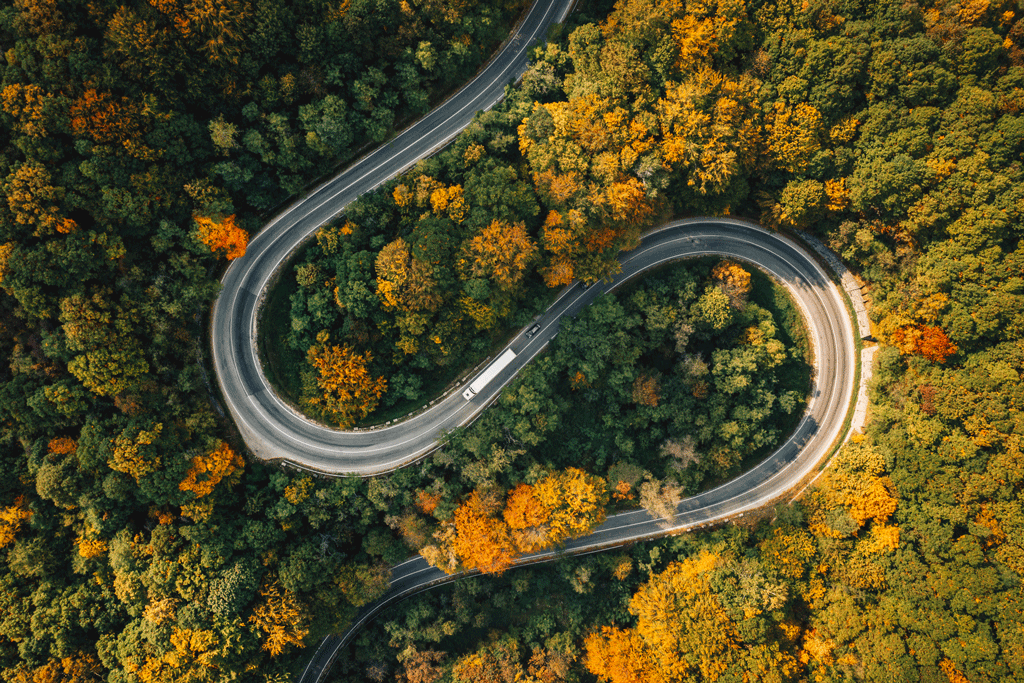 71f7737cc171a1fd5f2b640886829573 (1).png An electric truck driving on a winding road through fall trees